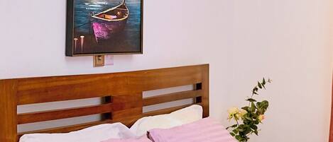 A hotel room with a wooden headboard, a bed with pink and white bedding, a framed picture of a beach scene, a bedside table with a phone, and a wall-mounted light fixture.
