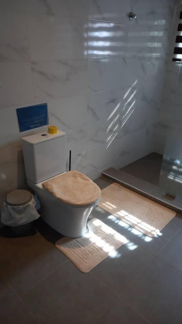 A bathroom with a toilet, a shower, and a mat. - JLK Guest House & Events Centre (Koforidua)