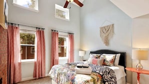 A bedroom with a large bed, two windows with red curtains, a ceiling fan, and a small table with a lamp. - Fells Point Fenced in Yard Weber Gas Grill (Baltimore)