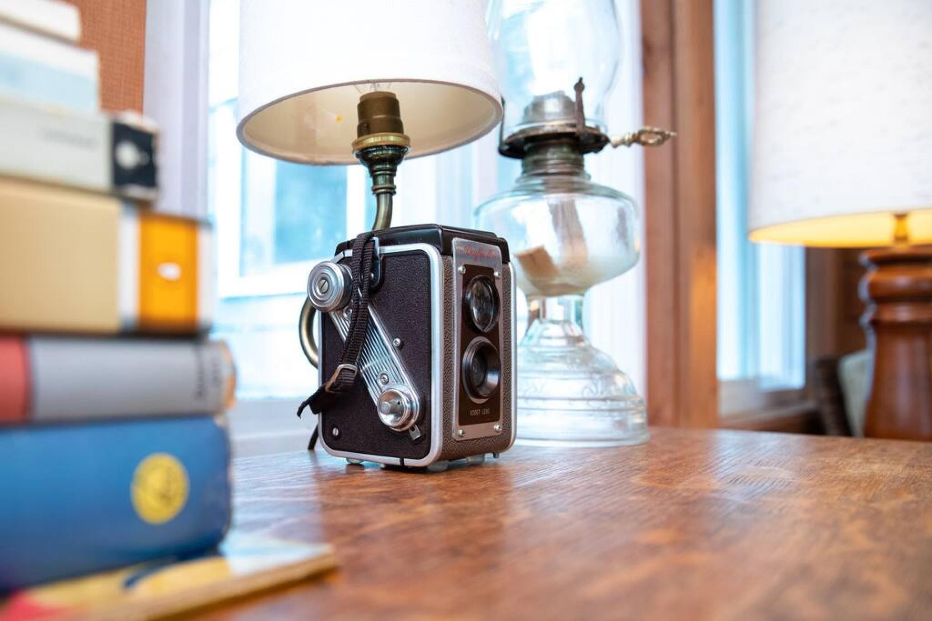 A vintage camera on a wooden table with books and a lamp in the background.