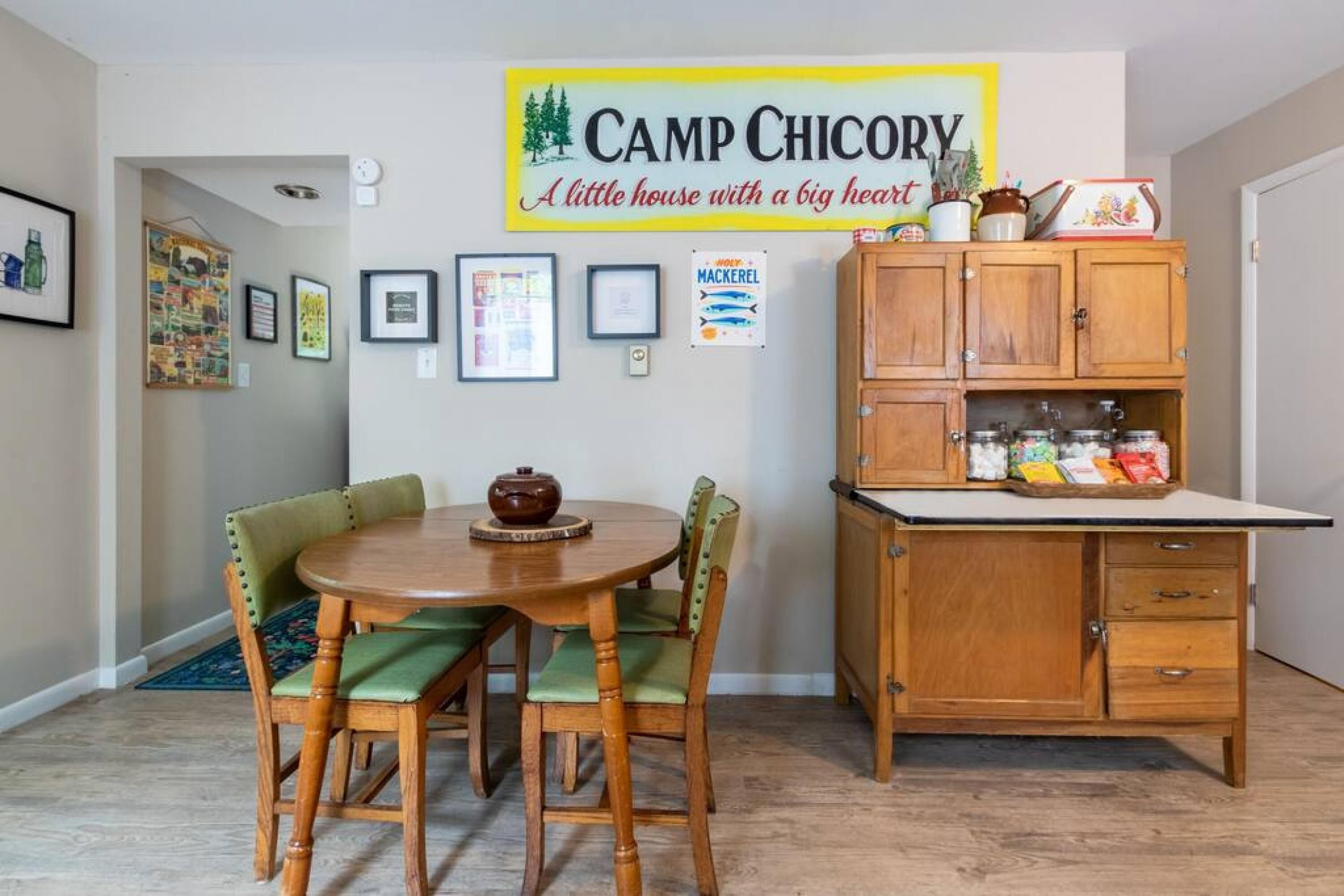 A dining area with a wooden table and chairs, a cabinet with kitchen items, and a sign above reading "Camp Chicory".