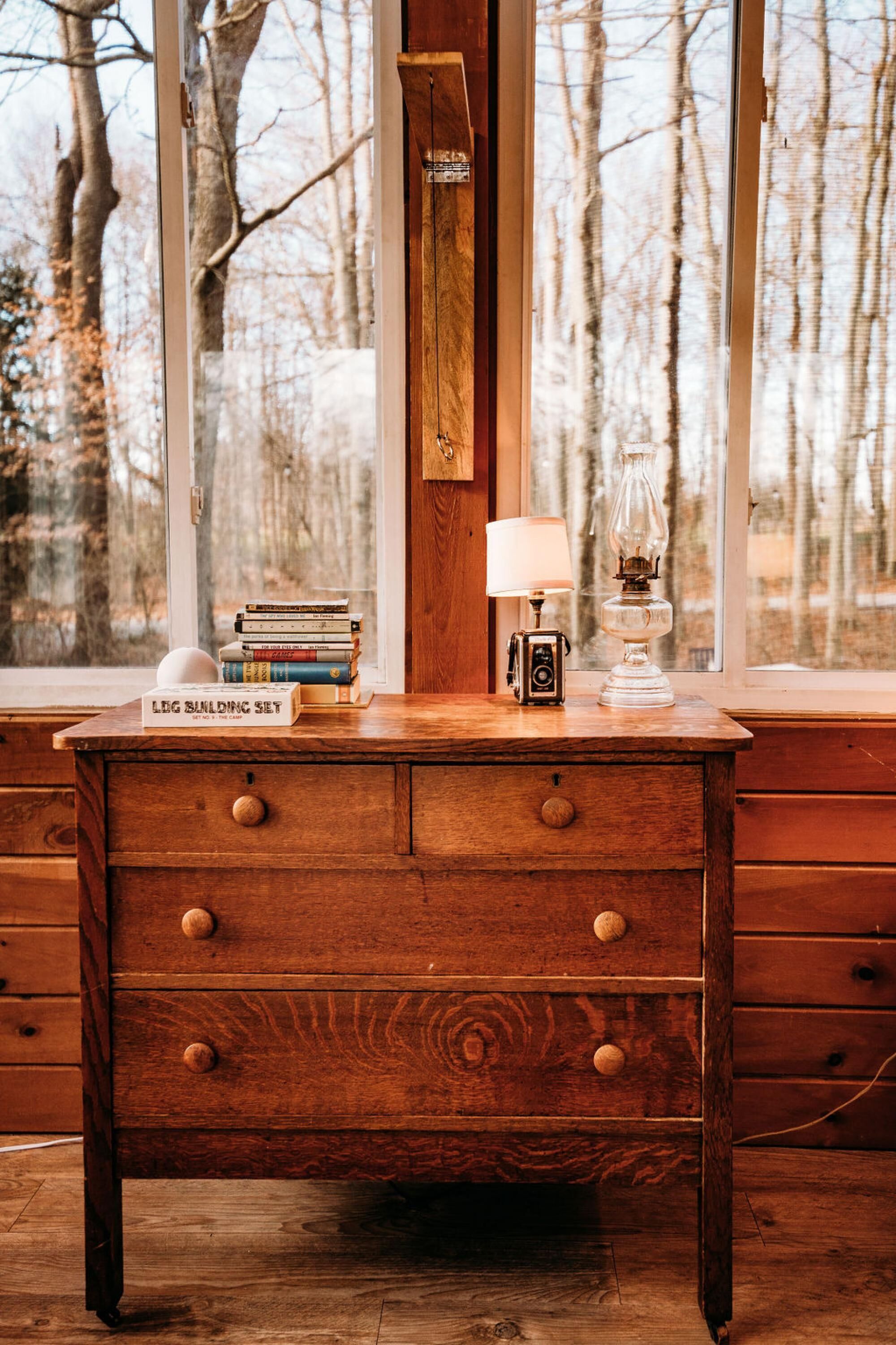 Un escritorio de madera con cajones, una pila de libros, una cámara y una lámpara junto a una ventana que da a vistas de árboles.