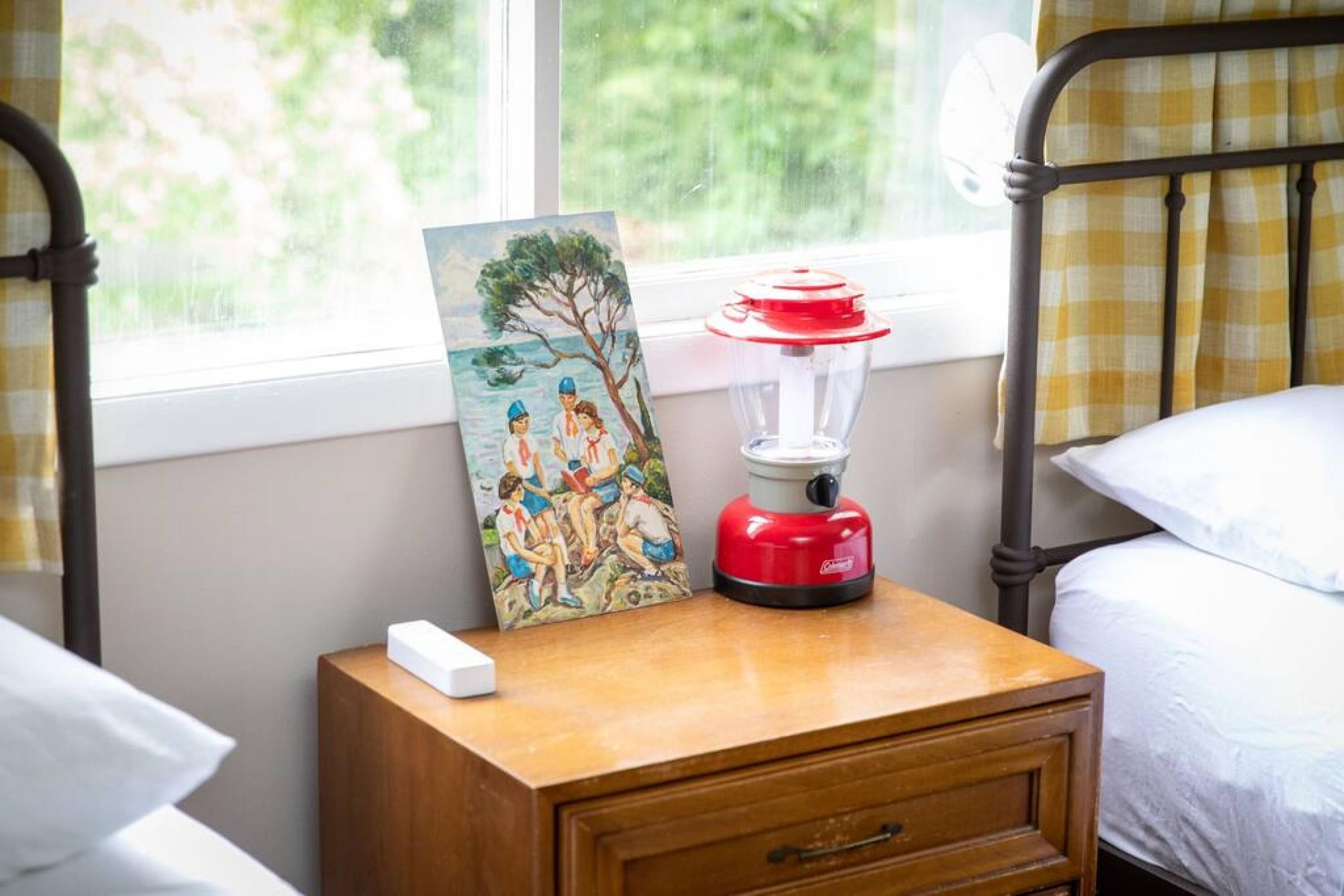 Une chambre à coucher avec une table de chevet, un tableau, une lampe rouge et blanche, et une fenêtre donnant sur des arbres.