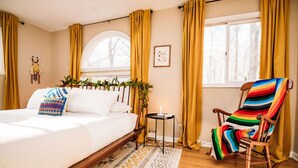 Une chambre à coucher avec un lit, un fauteuil à bascule, une petite table avec une bougie et une fenêtre avec des rideaux.