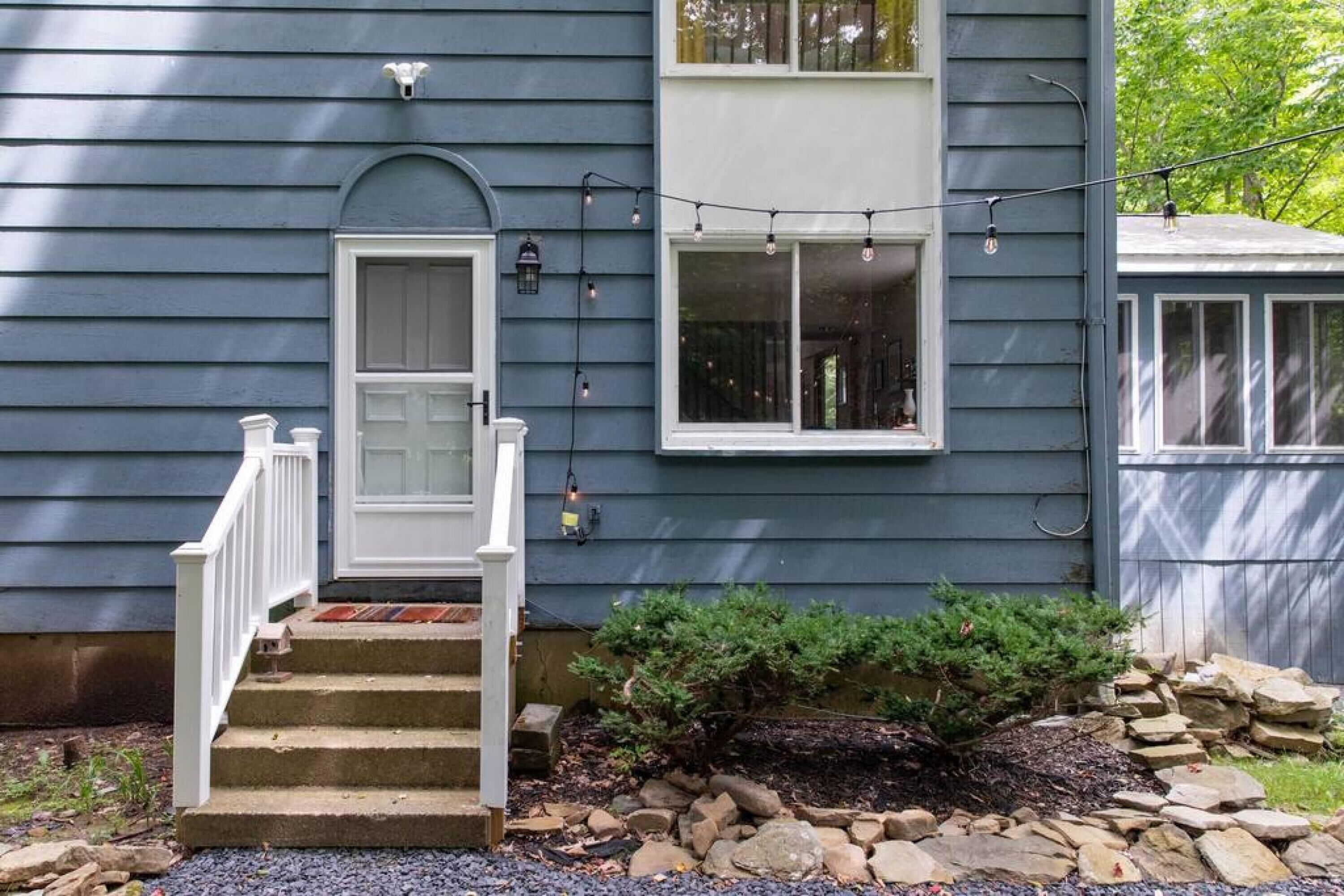 A house with a front porch, a small garden with rocks and a bush, and a string of lights.
