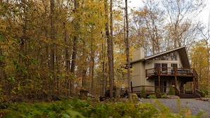 A cabin with a wooden deck and a staircase leading to the entrance, surrounded by a forest with autumn foliage.
