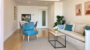 Een moderne woonkamer met een bank, een salontafel en een blauwe fauteuil. In de achtergrond bevindt zich een eethoek met een tafel en stoelen.