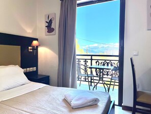 Habitación de hotel con cama, mesita de noche, lámpara, silla, mesa y balcón con vistas.