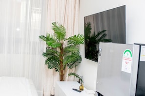 A modern room with a white bed, a large potted plant, a flat-screen TV, and a refrigerator.