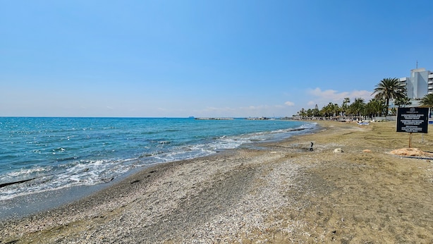 Una playa con piedras, un cartel que indica 'Sombrillas Reservadas para Huéspedes' y un cielo azul despejado.