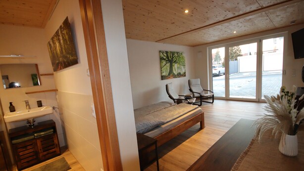 A bedroom with a wooden bed, a bench, a chair, a small table, a sink, and a painting on the wall.
