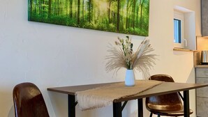 A dining area with a wooden table, two leather chairs, a vase with dried plants, and a large forest-themed painting on the wall.