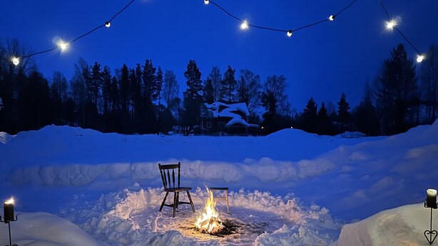 A snow-covered outdoor area with a fire pit, a chair, and string lights.