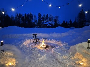 Lumipeitteinen ulkoalue, jossa on nuotiopaikka, tuoli ja valoketju.