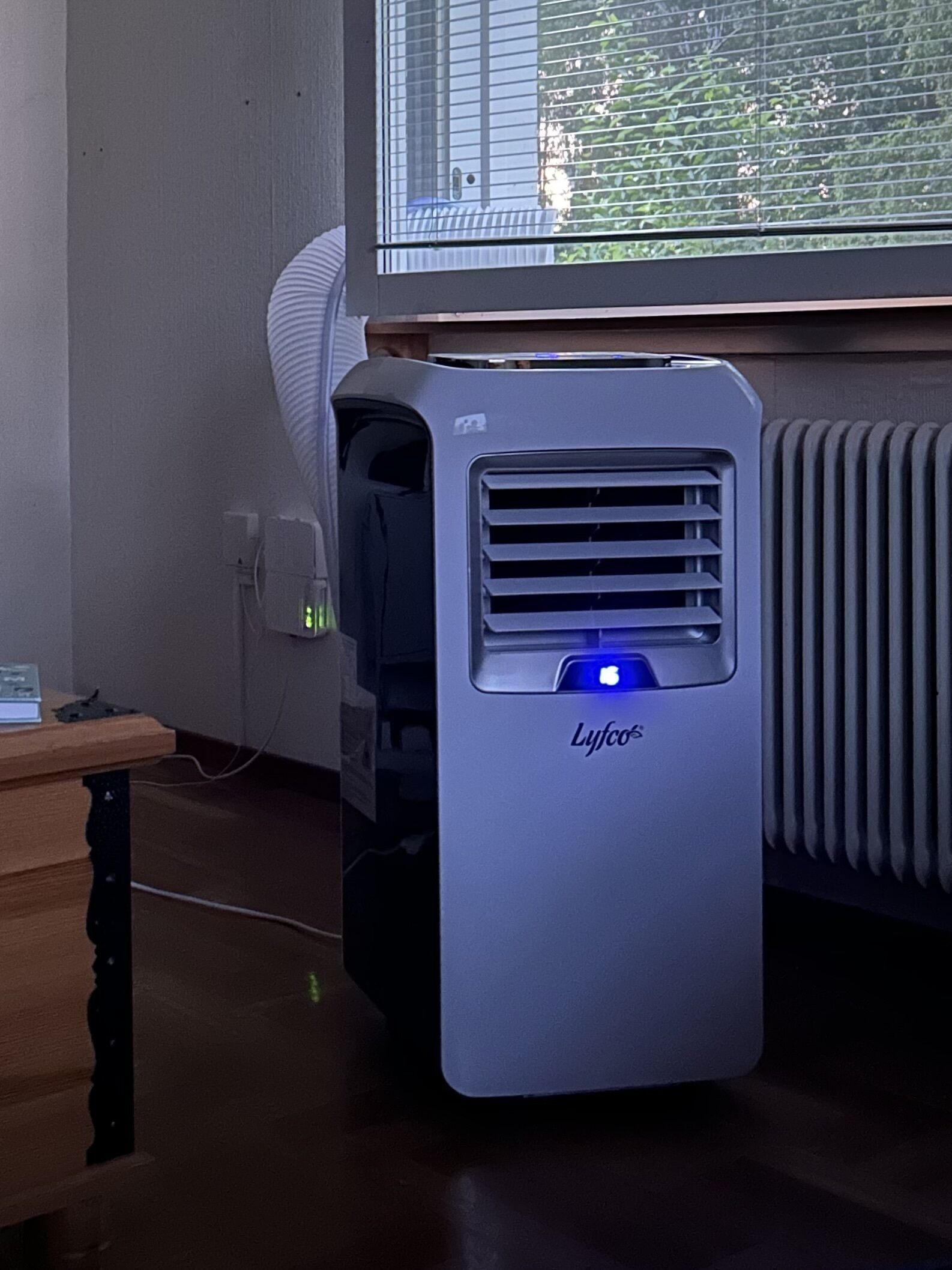 Un climatiseur Lyco dans une pièce équipée d’une fenêtre, d’un radiateur et d’un bureau en bois.