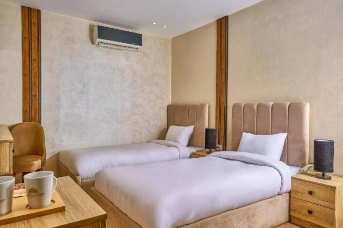 Quarto de hotel com duas camas, uma mesa de cabeceira de madeira, uma mesa com duas canecas e um ar condicionado fixado na parede.