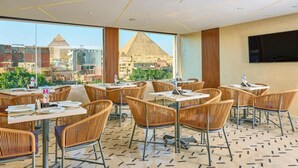 A restaurant with wicker chairs and tables, large windows with a view of pyramids, and a flat-screen TV on the wall. - Cleopatra Tower Pyramids Hotel (Giza)