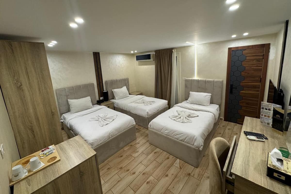 Quarto de hotel com três camas, uma escrivaninha de madeira, uma cadeira, uma mesinha e uma televisão.