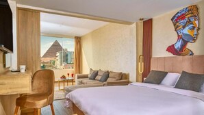 A hotel room with a bed, a desk, a chair, a couch, and a large window offering a view of a pyramid. - Cleopatra Tower Pyramids Hotel (Giza)