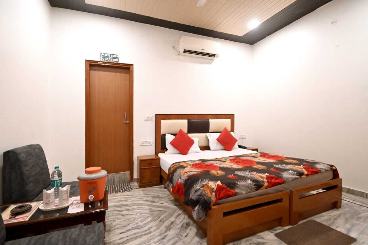 A hotel room with a bed, a nightstand, a chair, a table with a water bottle, a trash bin, and an air conditioning unit.