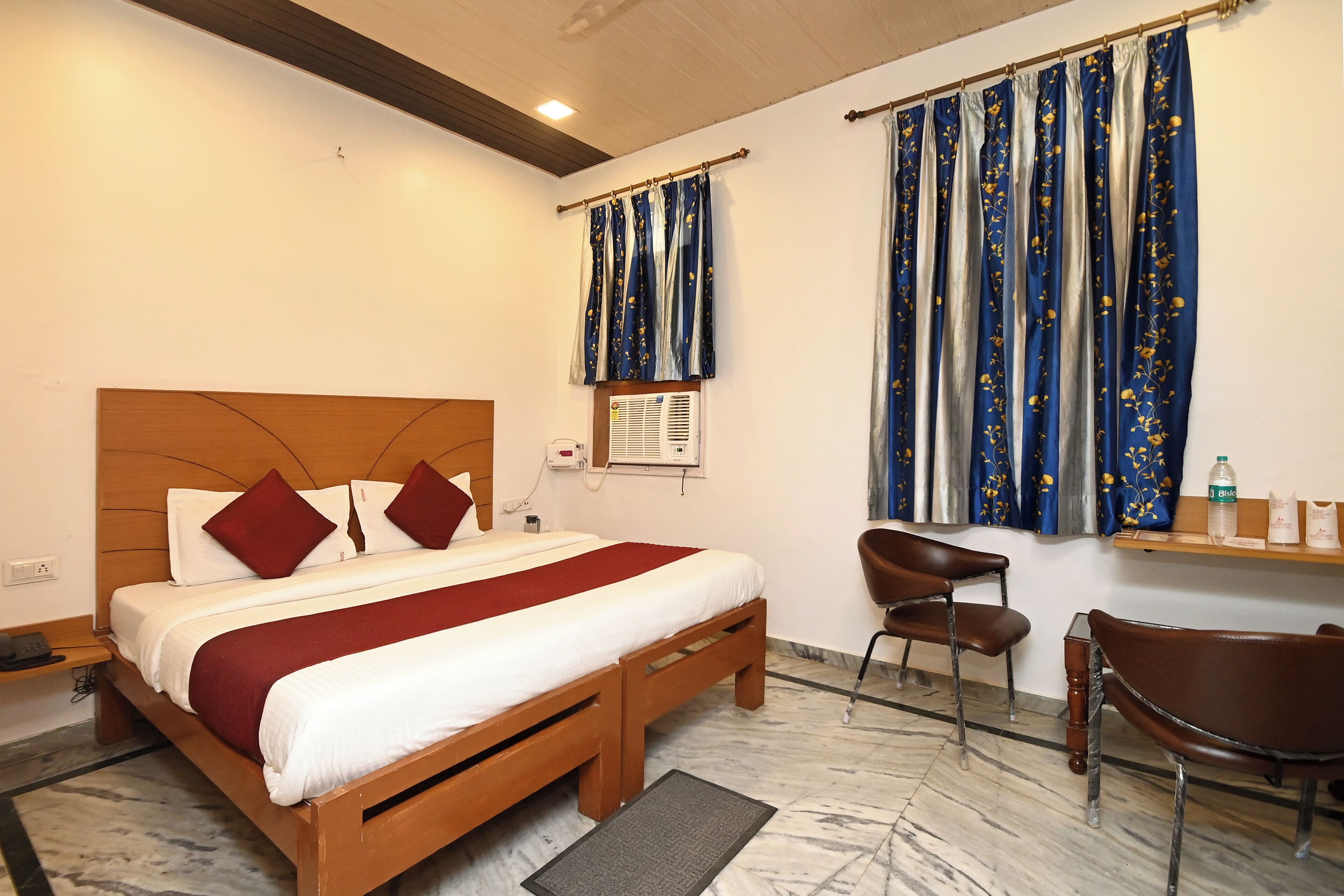 A hotel room with a wooden bed, red and white bedding, a marble floor, a desk with a chair, and blue curtains with a floral pattern.