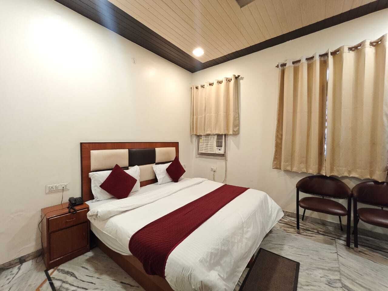 A hotel room with a bed, a nightstand, a chair, a window with curtains, and a ceiling with wooden beams.