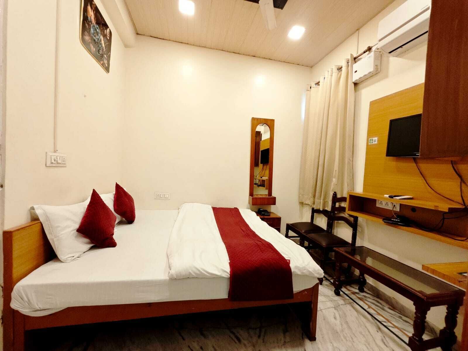 A hotel room with a bed, a bedside table, a mirror, a TV mounted on the wall, and a wooden bench.