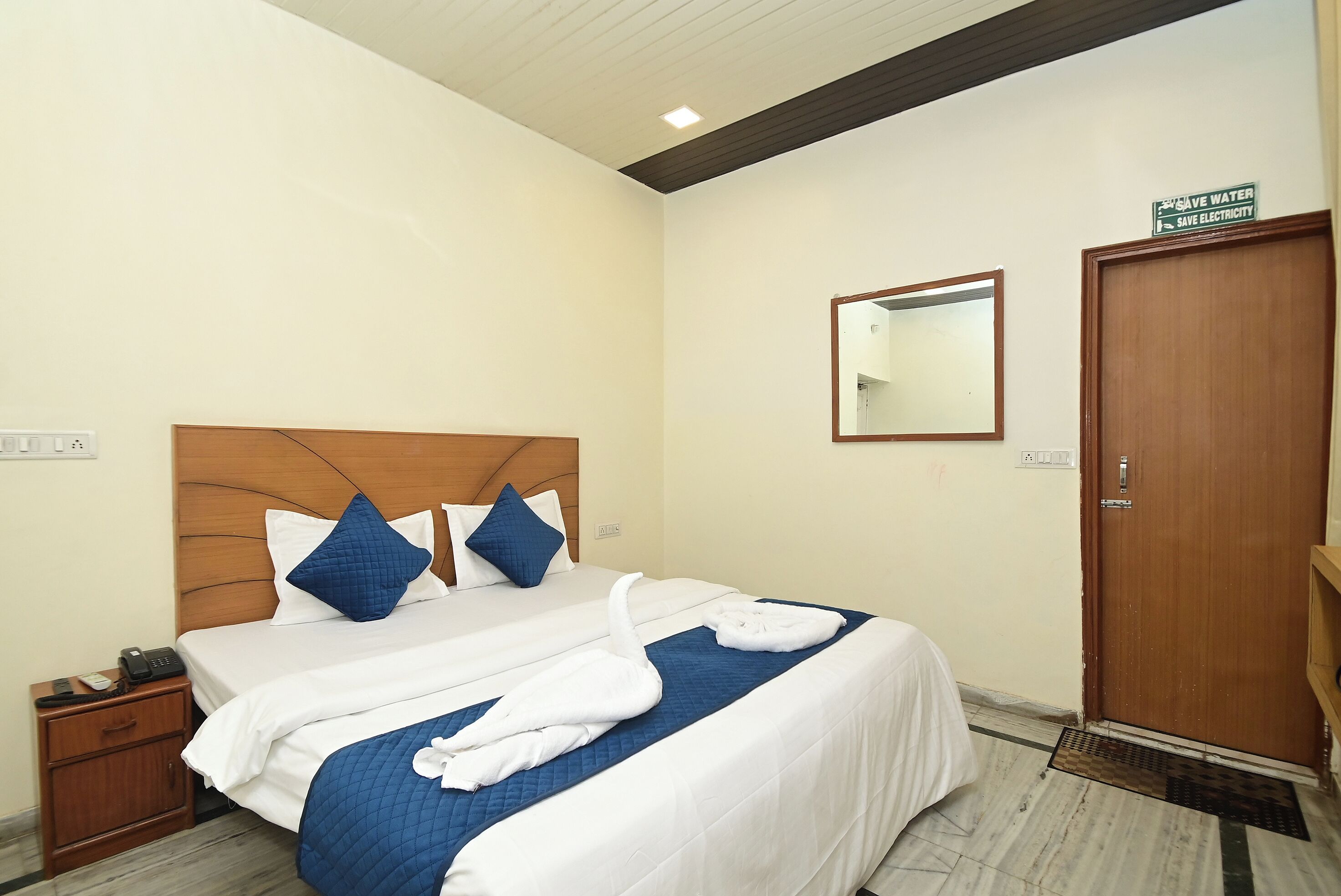 Hotelkamer met houten hoofdbord, bed met wit en blauw beddengoed, nachtkastje met telefoon, spiegel aan de muur en deur met bordje.