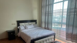 A bedroom with a bed, a nightstand, a window with curtains, and a view of the outside. - UNIQUE GUEST HOUSE (Addis Ababa)