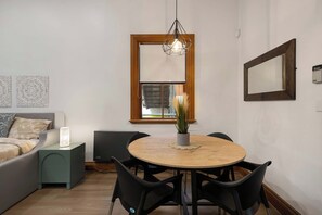 A modern living room with a round wooden dining table, black chairs, a grey sofa, a small side table with a lamp, a decorative wall piece, a large window with a view, and a hanging geometric light fixture. - Lambton Quay Loft - Sleeps 10 (Wellington)