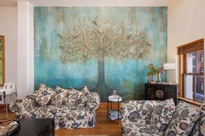 Ein Wohnzimmer mit zwei gemusterten Sofas, einem großen Blumenbild an der Wand, einem kleinen Beistelltisch mit einer Vase und einem Fenster mit Jalousien.