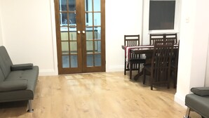 Ein geräumiges Wohnzimmer mit Holzboden, einem grünen Sofa, einem Esstisch mit Stühlen und einer Glastür zum Balkon.