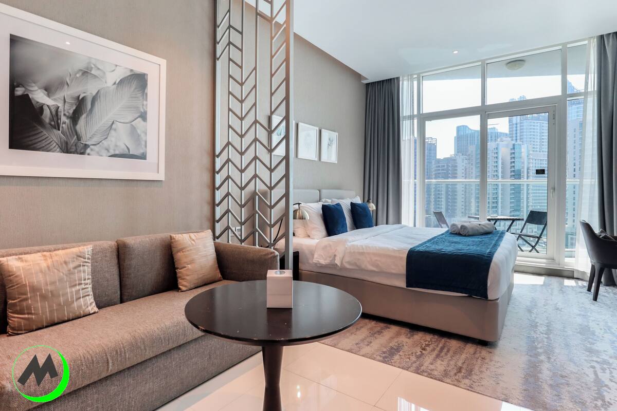 A modern hotel room with a sofa, a bed, a small table, and a city view through large windows.
