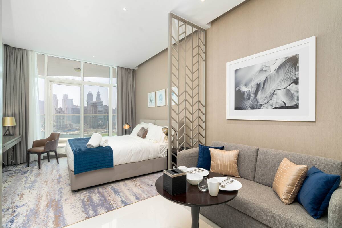 A hotel room with a bed, a sofa, a dining table, and a large window with a city view.