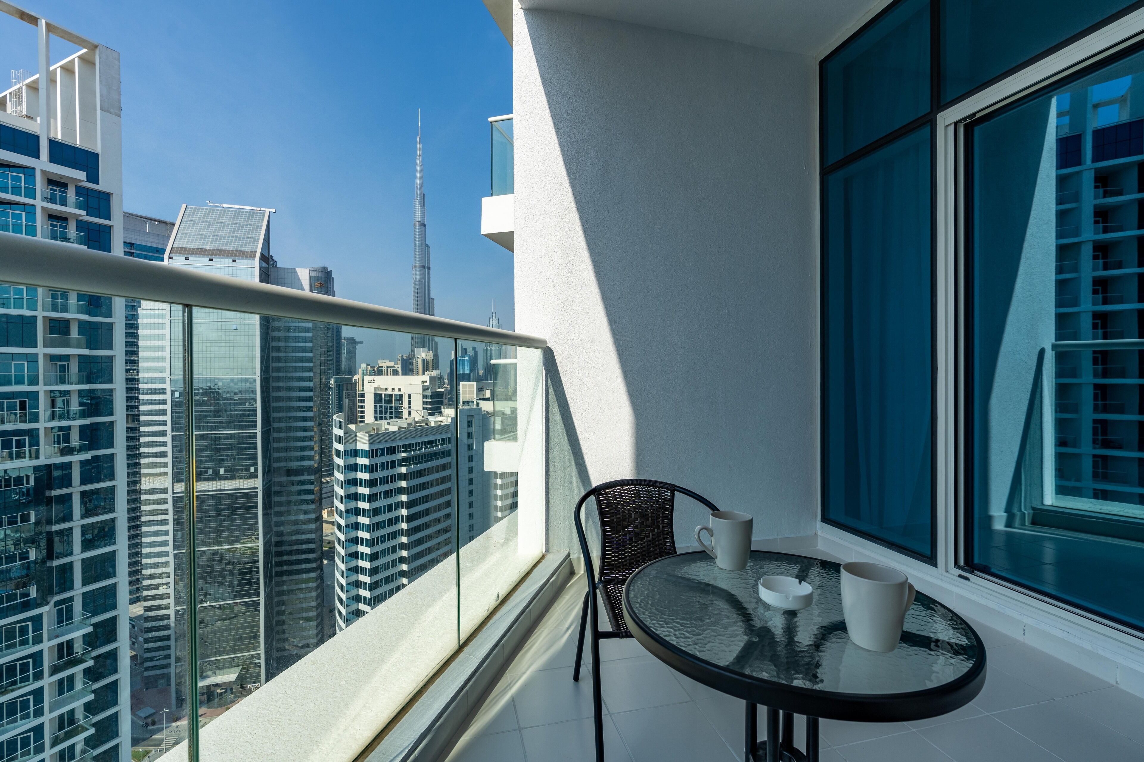 A balcony with a glass railing, a table with two white cups, and a view of a cityscape including a tall skyscraper.