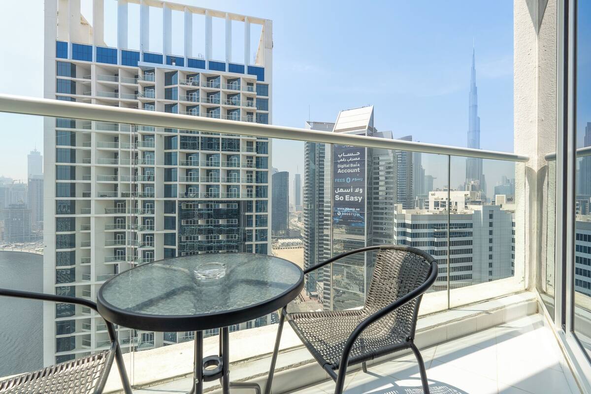 A balcony with a glass table and two chairs, overlooking a cityscape with modern buildings and the Burj Khalifa.