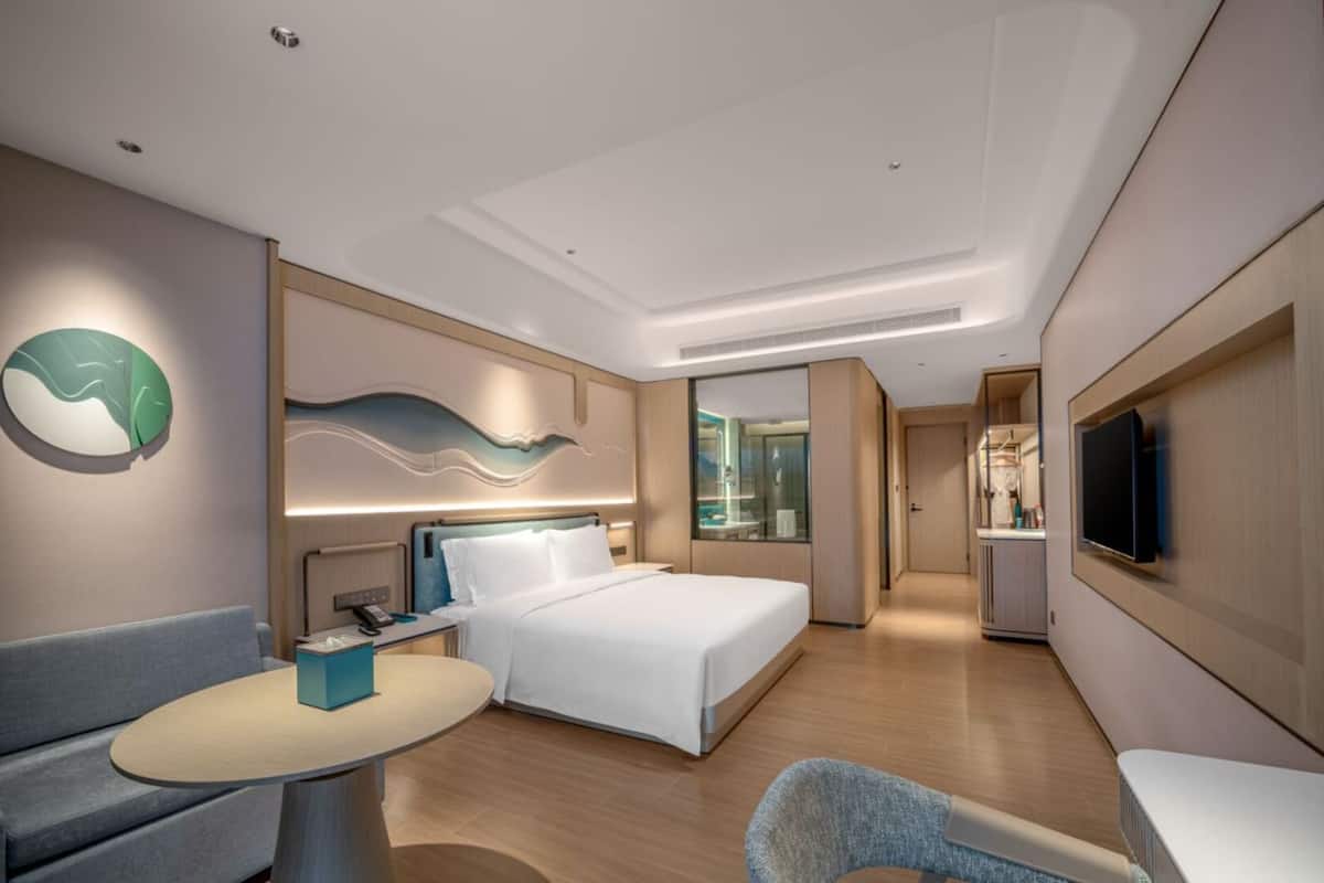 A modern hotel room with a bed, a desk, a chair, a TV, and a bathroom visible through a glass partition.