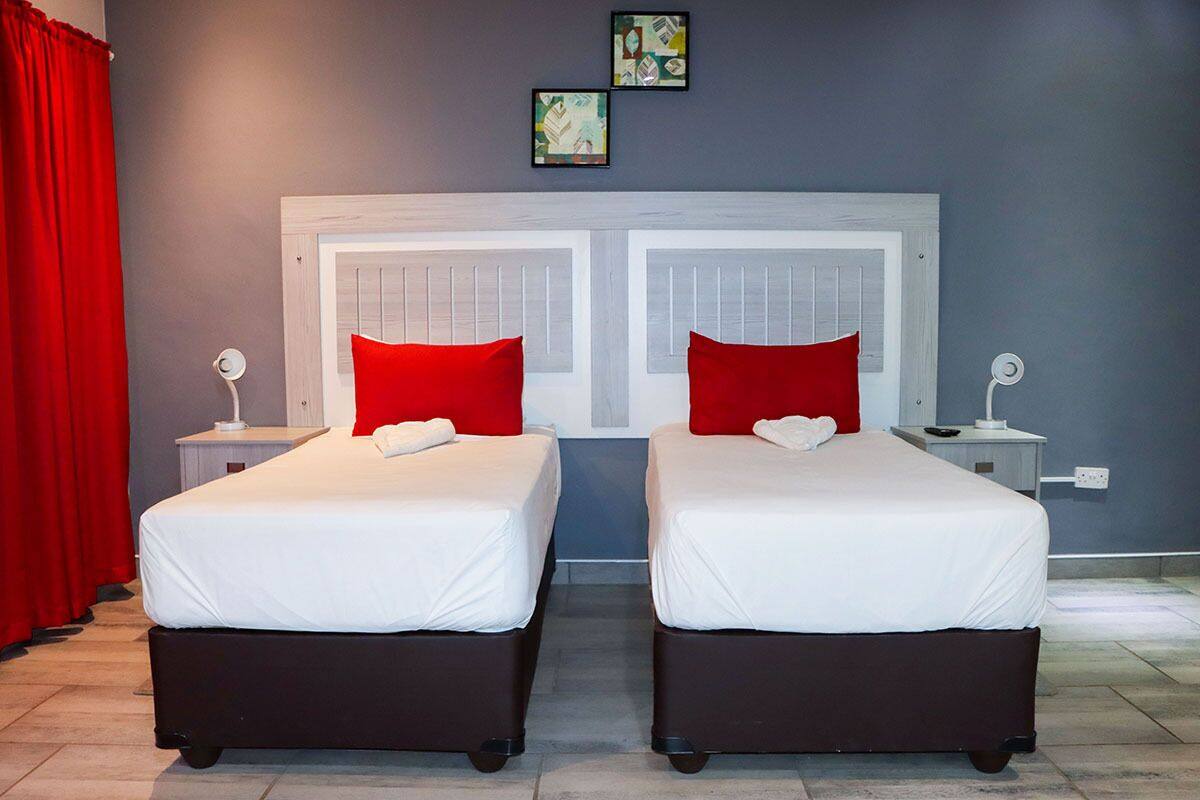 Une chambre double avec deux lits, chacun doté d’un matelas blanc et de coussins rouges, le tout adossé à un mur gris orné de deux tableaux encadrés.