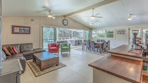 A spacious living area with a large sofa, a coffee table, and a dining table with chairs. There are ceiling fans and a clock on the wall.