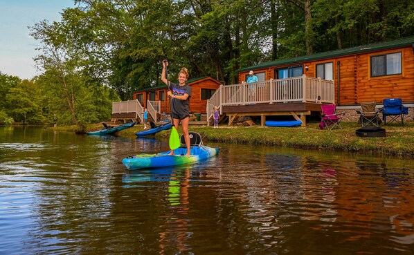 A wooden cabin with a deck, kayaks, and a paddleboarder.