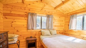 Une chambre dans une cabane en bois, comprenant un lit, une table de chevet, une commode et deux fenêtres avec des rideaux.