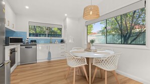 Uma cozinha moderna com armários brancos, uma área de jantar com mesa redonda e cadeiras brancas, e uma grande janela que oferece vista para árvores e um edifício com telhado vermelho.