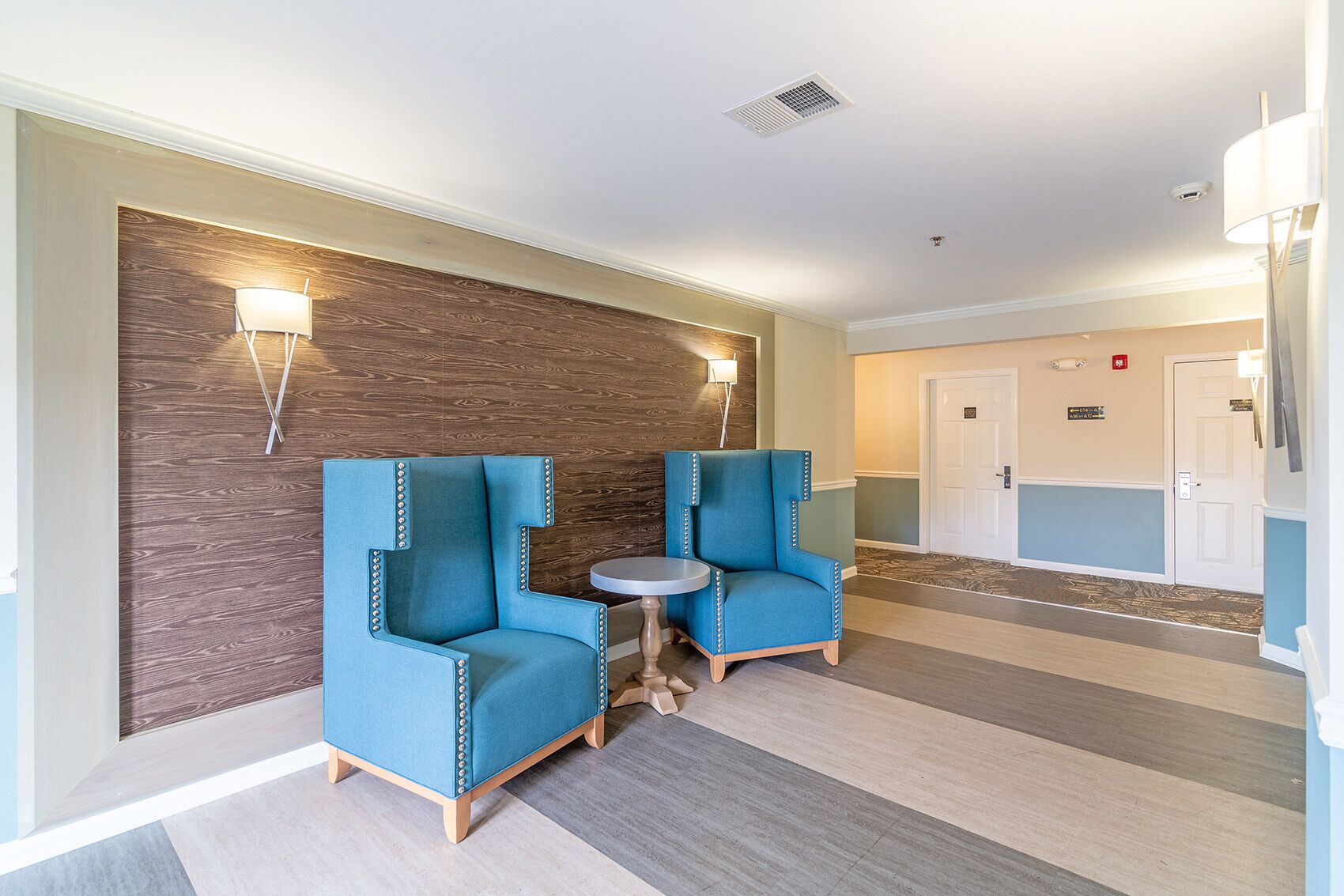 Un vestíbulo de hotel con dos sillones azules, una pequeña mesa redonda y una pared de acento de madera.