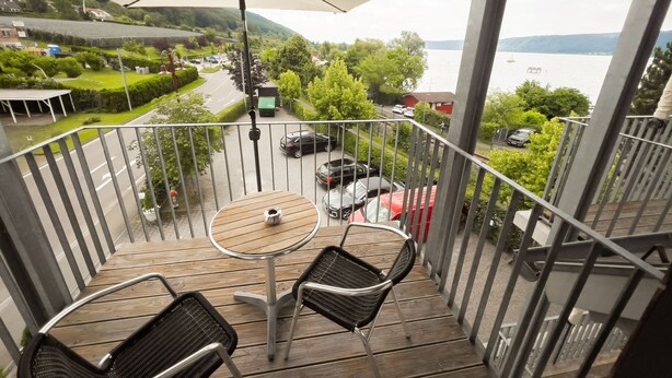 Een balkon met een tafel en stoelen, een grote parasol en uitzicht op een meer en groen.