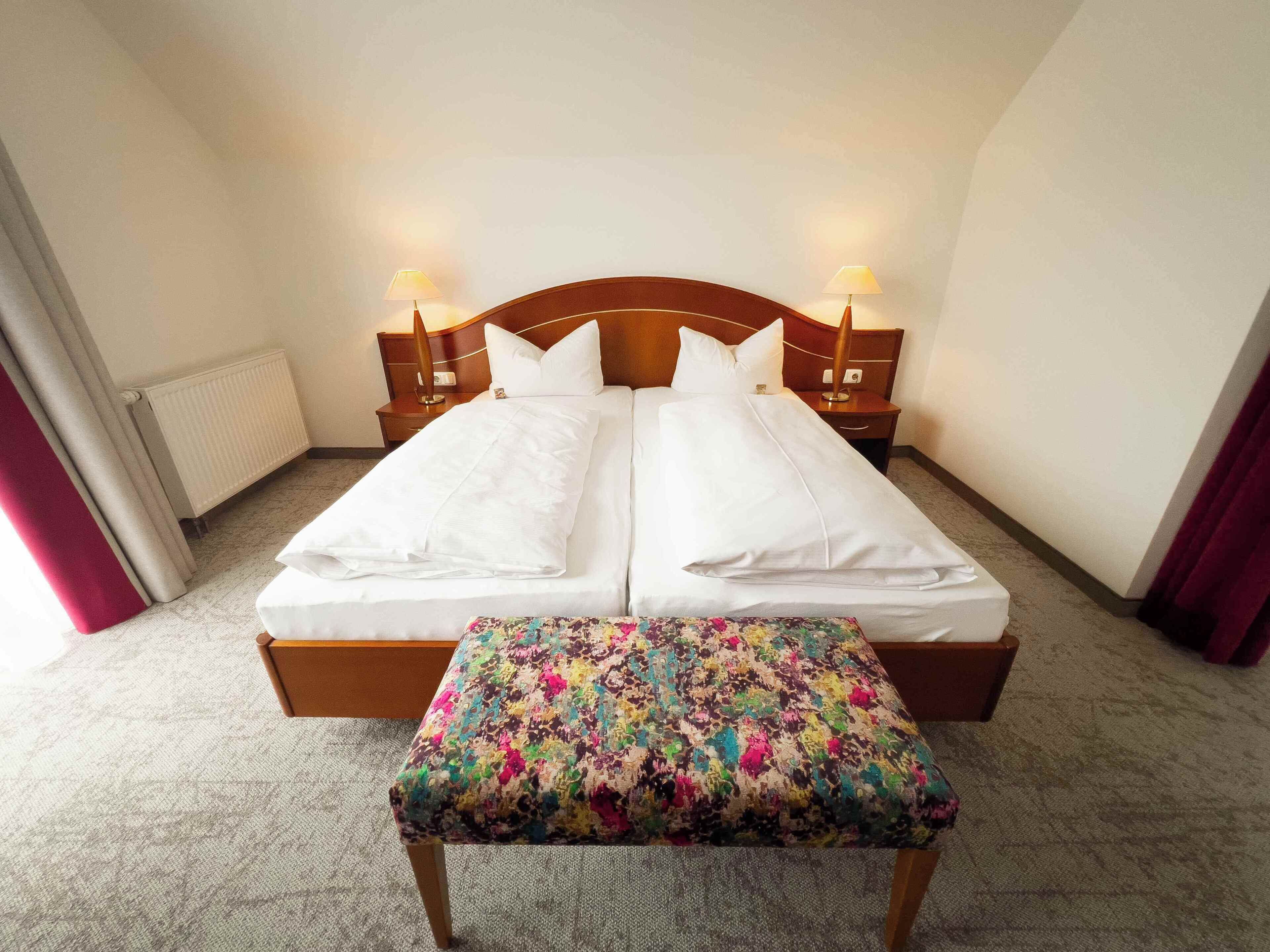 A double bed with white linens, two bedside tables with lamps, a radiator, and a colorful bench.