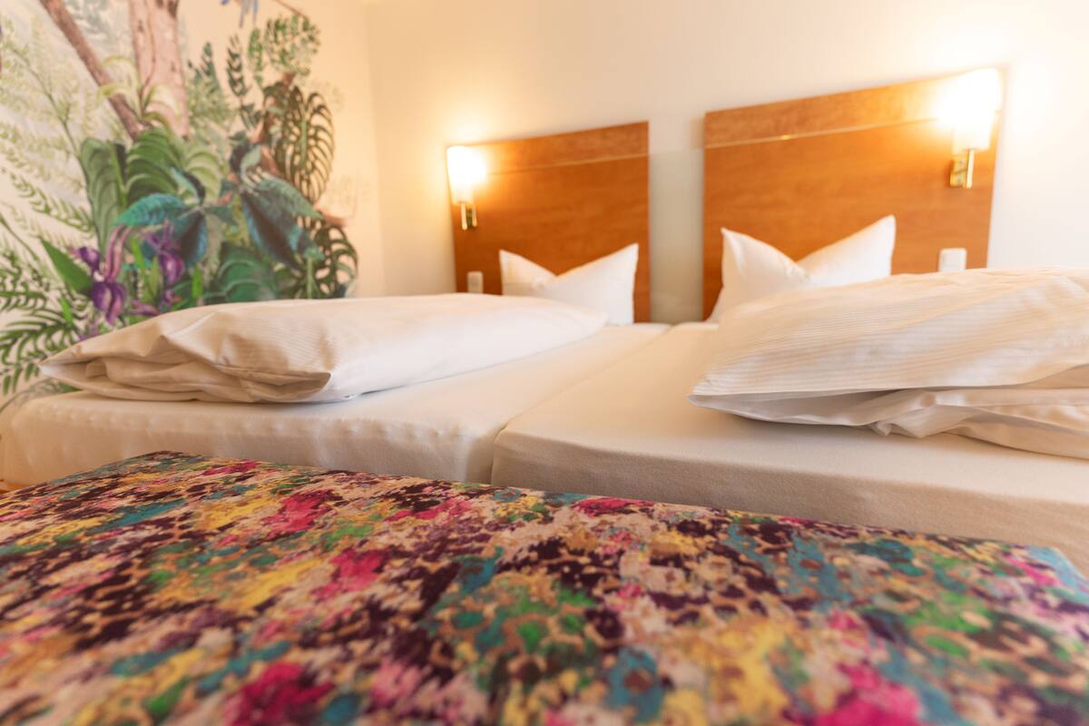 Een hotelkamer met twee bedden, elk voorzien van witte lakens en kussens, en een kleurrijke bloemenprint als dekbed.