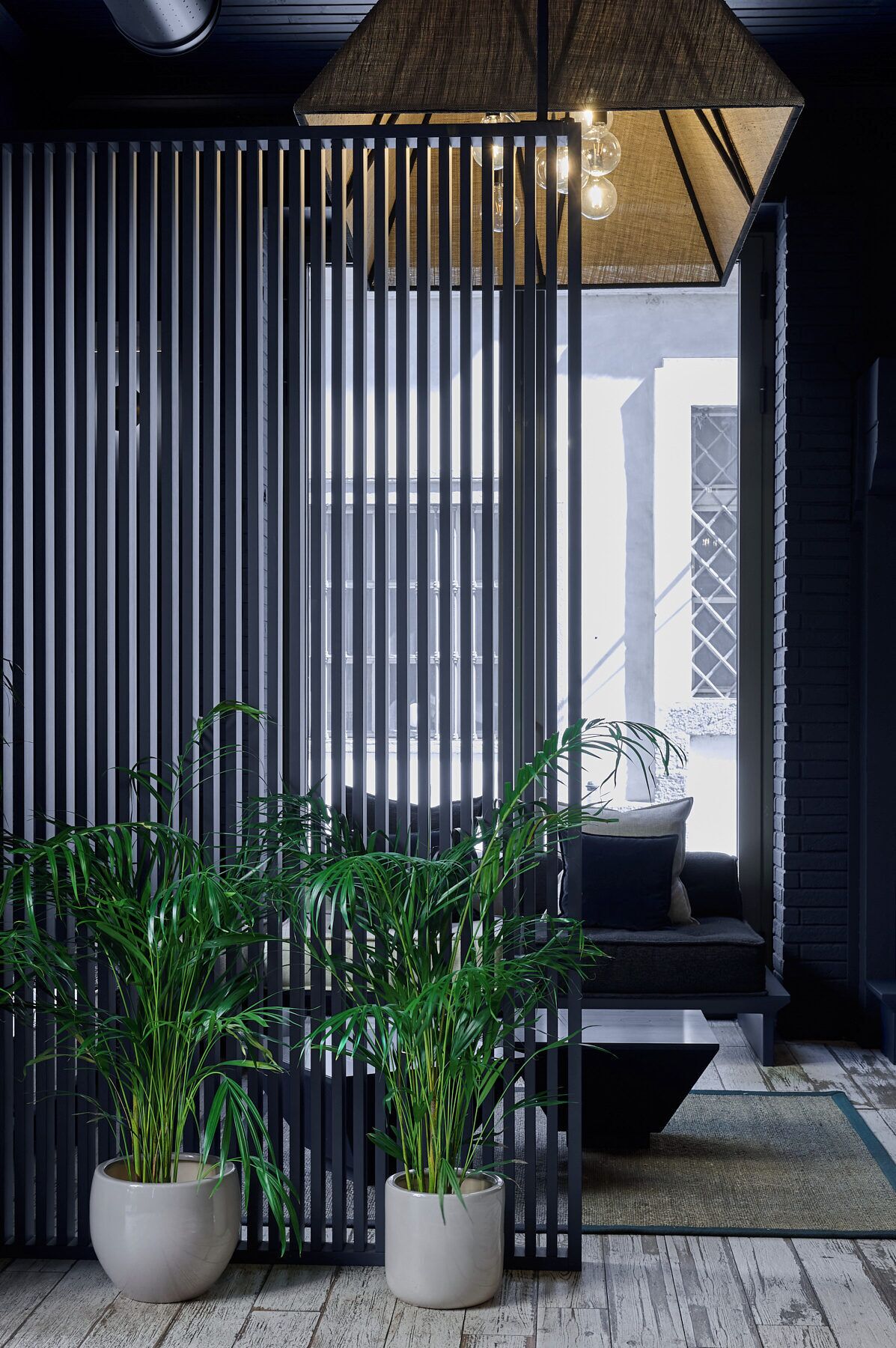 Un interior moderno con un separador de listones de madera verticales, un banco con cojines y dos plantas en macetas.