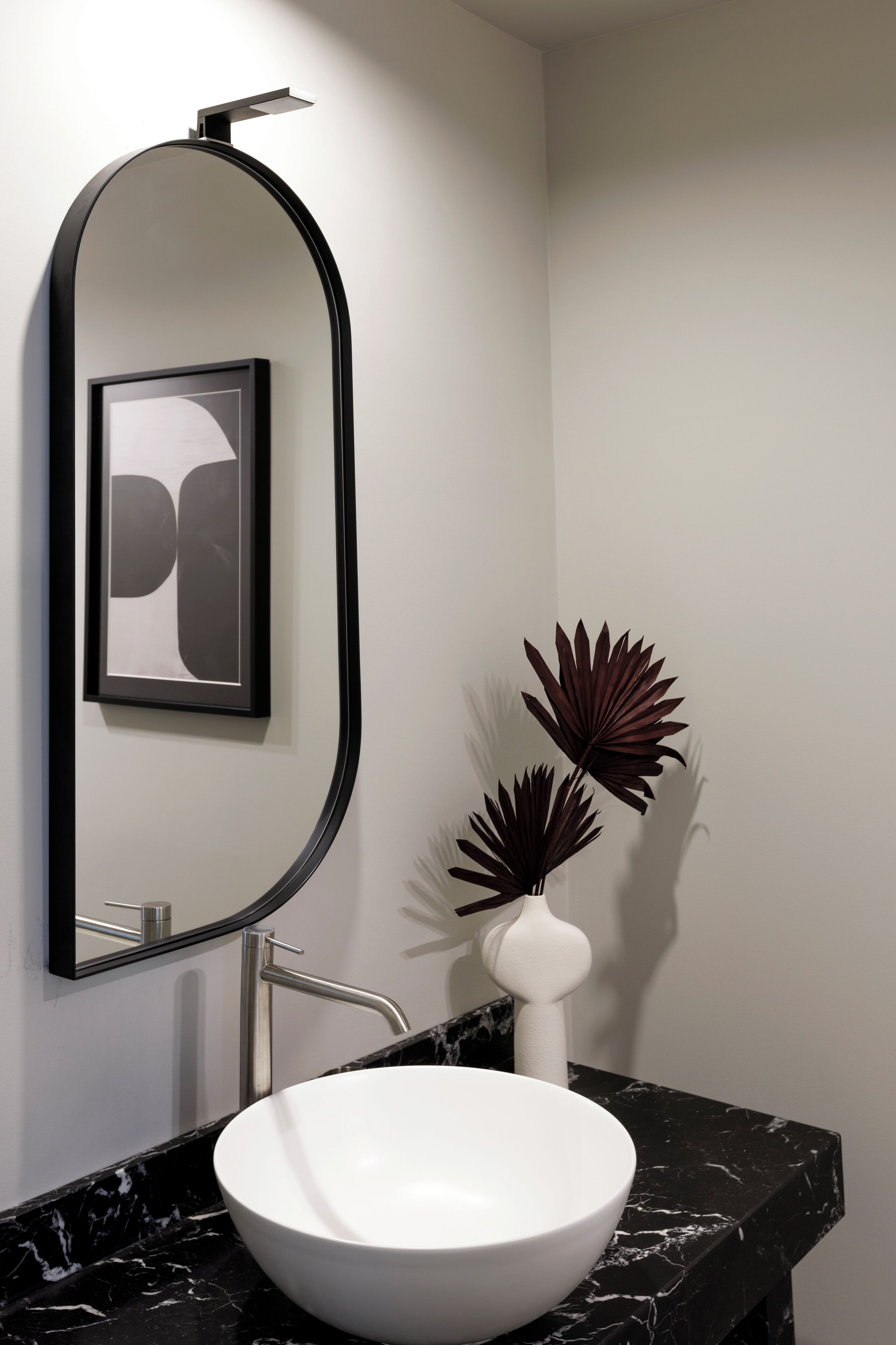 A modern bathroom with a white sink, black marble countertop, a large mirror, and decorative plants.