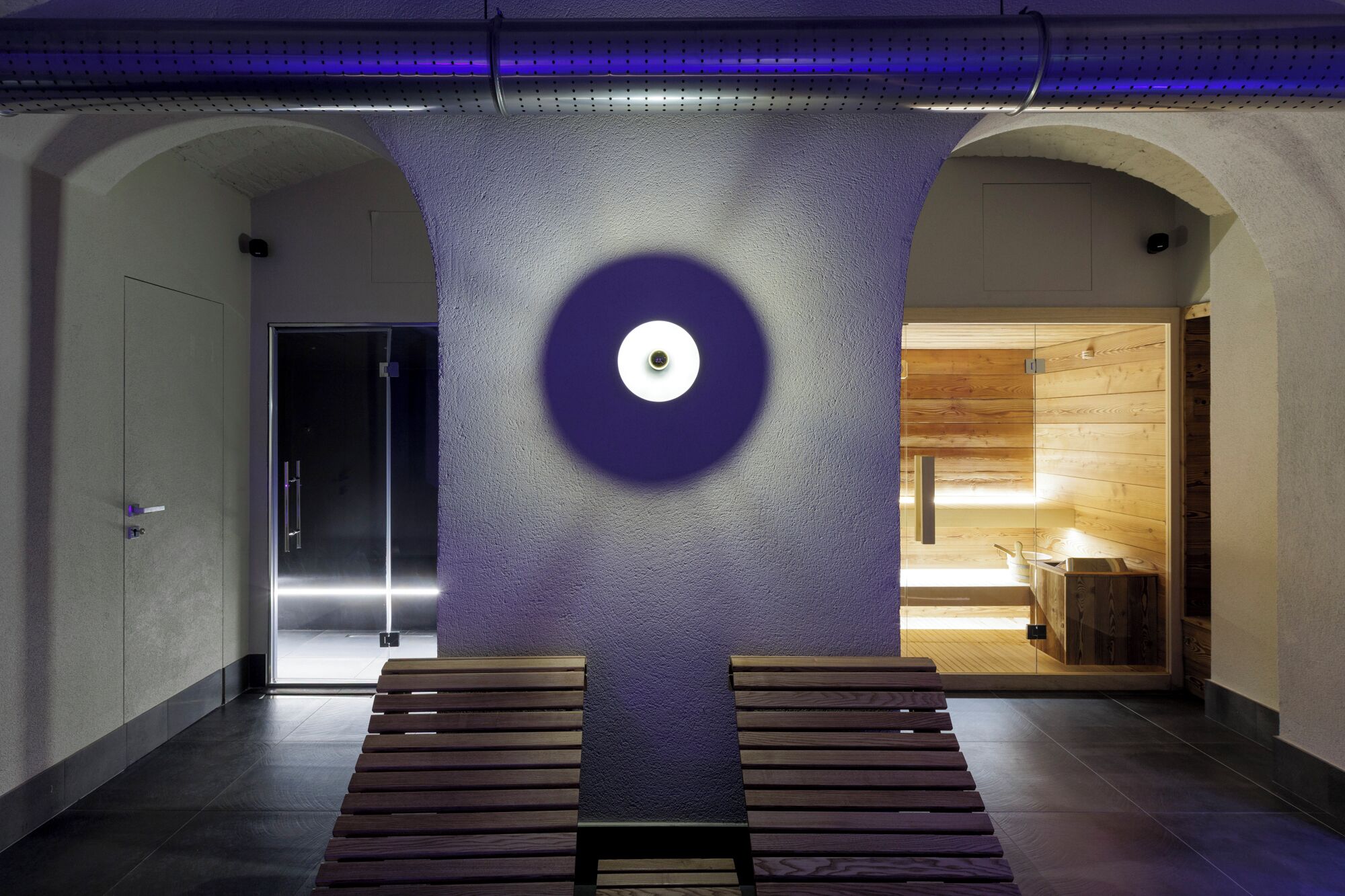 A modern interior with a central circular light fixture, wooden benches, and a visible sauna area.