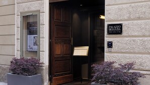 Entrada de un hotel con una puerta de madera grande, un cartel que dice "PALAZZO BELLEZIA" y dos macetas con follaje púrpura.
